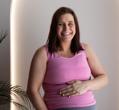 Pregnant woman wearing a pink feeding tank top and grey bump friendly skirt, standing against a neutral background.